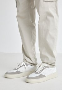White and gray sneakers with a textured suede accent, featuring perforations and flat laces, paired with light beige pants.
