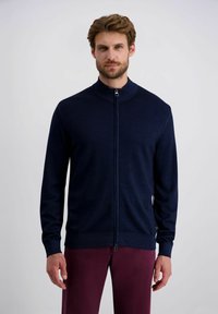 Marineblaue Reißverschlussstrickjacke mit hohem Kragen, gerippten Bündchen und Saum, glatter Textur, minimalistisches Design, getragen über kastanienbraunen Hosen.