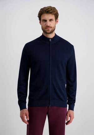 Cardigan zippé bleu marine avec un col montant, des poignets et un ourlet côtelés, texture lisse, design minimaliste, porté sur un pantalon bordeaux.