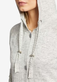 Gros plan sur une personne portant un sweat à capuche gris clair avec fermeture à boutons et cordons de serrage dotés de glissières cylindriques en argent.