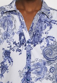 Dettaglio di una camicia bianca con motivi floreali e paisley blu, con bottoni blu e colletto aperto su pelle scura.
