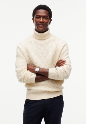 Hombre con los brazos cruzados, vestido con un jersey de cuello alto de punto trenzado color crema y pantalones oscuros, sonriendo suavemente sobre un fondo liso.