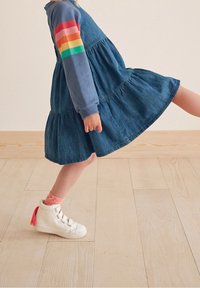 Abito in denim con un design a strati, maniche lunghe di colore blu con un motivo a strisce arcobaleno. Sneakers alte bianche con dettagli rosa.
