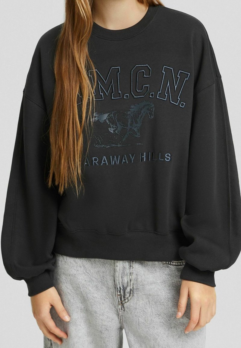 Personne portant un sweat-shirt noir à manches bouffantes avec le texte "M.C.N. Faraway Hills" et un graphique de cheval, associé à un jean clair.
