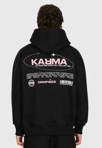 Personne portant un sweat à capuche noir avec le texte rose "KARMA" et un texte blanc plus petit sur le karma, ainsi que "DROPSIZE" et un design de code-barres au dos.