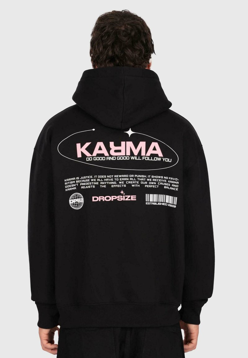 Personne portant un sweat à capuche noir avec le texte rose "KARMA" et un texte blanc plus petit sur le karma, ainsi que "DROPSIZE" et un design de code-barres au dos.