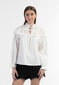 Blusa bianca in cotone con colletto arricciato, chiusura con laccetto e maniche lunghe con polsini elasticizzati. Abbinata a pantaloni corti di denim neri.