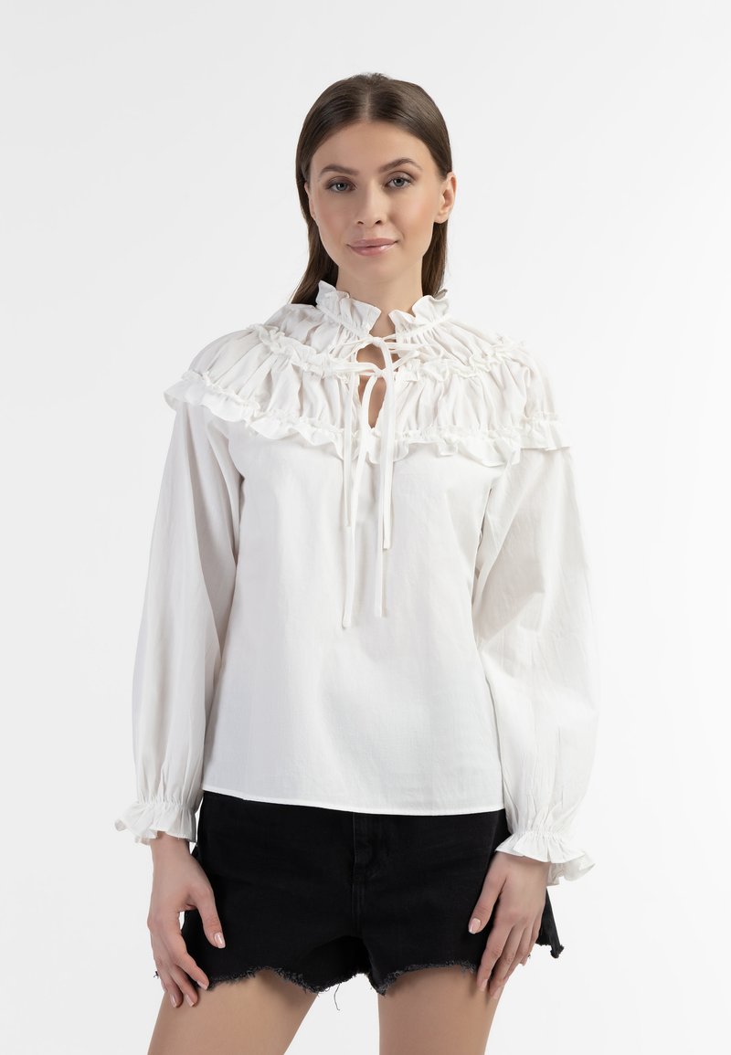 Blusa bianca in cotone con colletto arricciato, chiusura con laccetto e maniche lunghe con polsini elasticizzati. Abbinata a pantaloni corti di denim neri.