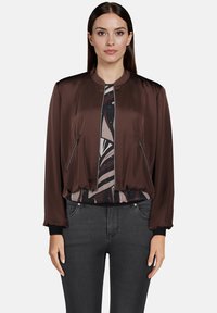 Brun satin bomberjakke med en tætsiddende design, ribbede manchetter og to sidelommer med lynlås, lagt over en mønstret skjorte. Sorte jeans bæres.