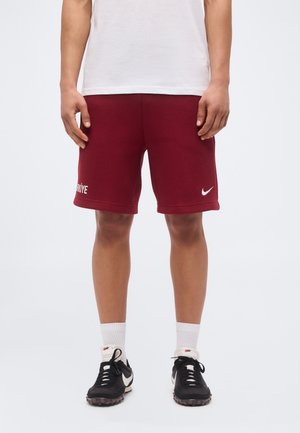 Personne portant un short Nike bordeaux avec le texte « Türkiye », des chaussettes blanches montantes et des baskets Nike noires, debout sur un fond uni.