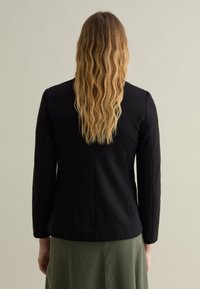 Blazer negro ajustado con mangas largas, hombros estructurados y detalles de costura sutiles, usado sobre una falda verde. Textura suave.