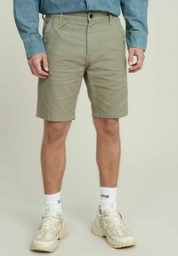 Shorts verde oliva realizzati in tessuto misto cotone, con tasche frontali e una finitura pulita. Indossati con sneaker beige e calzini bianchi.