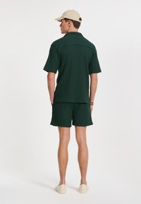 Cămașă verde texturată cu mânecă scurtă și pantaloni scurți asortați, confecționată dintr-un material ușor. O șapcă bej și pantofi de culoare deschisă completează ținuta.