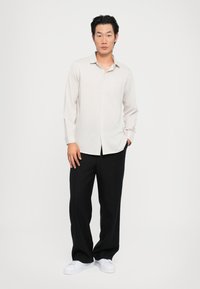 Lys beige langermet skjorte med krage som kan knytes, kombinert med svarte vide bukser og hvite sneakers. Jevn stofftekstur.