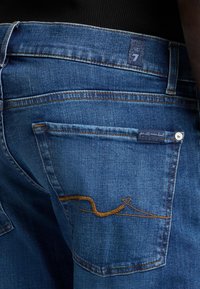 Blå denimjeans med en bakficka som har gul sömnad i ett vågmönster. Inkluderar en tygetikett nära midjan.