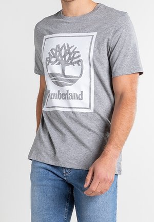 Homme portant un t-shirt gris Timberland avec logo arbre et un jean bleu, debout devant un fond clair uni.
