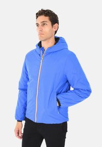 Uomo che indossa una giacca con cappuccio di un brillante blu, con cerniera frontale, in piedi con una mano nella tasca su uno sfondo chiaro e uniforme.