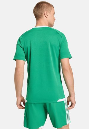 Mężczyzna stojący tyłem, ubrany w zielony sportowy t-shirt z krótkim rękawem oraz dopasowane krótkie spodenki z białymi paskami po bokach.