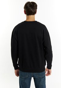Felpa nera con scollatura rotonda, maniche lunghe e polsini a coste. Tessuto morbido con una vestibilità rilassata, indossata con jeans blu.