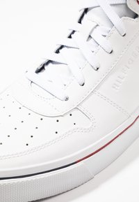 Bílé tenisky s perforovanou špičkou, plochými tkaničkami a značkovou stranou. Mají gumovou podrážku s barevným akcentem. Hladká kožená textura.