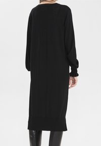 Robe noire à manches longues avec une coupe décontractée, présentant une texture côtelée et des poignets volantés, atteignant une longueur mi-mollet avec un ourlet droit.