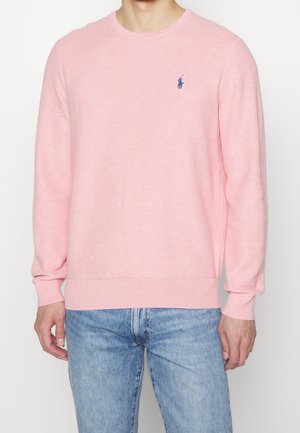 Mann trägt einen hellrosa Pullover mit langen Ärmeln und einem kleinen blauen gestickten Logo auf der Brust, kombiniert mit hellblauen Jeans.