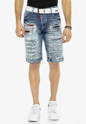 Blaue, zerstörte Denim-Shorts mit mehreren Rissen, kombiniert mit einem weißen Nieten-Gürtel. Getragen mit weißen Sneakers. Vorderansicht.
