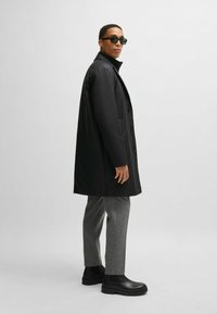 Cappotto imbottito nero con colletto alto e chiusura a bottoni, abbinato a pantaloni grigi e stivaletti neri. Il tessuto ha una texture liscia.