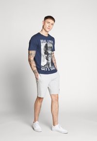 Camiseta de marina con un estampado gráfico que dice "VIDAS DUALES" en blanco, combinada con pantalones cortos grises claros. El modelo lleva zapatillas blancas; tatuajes visibles.