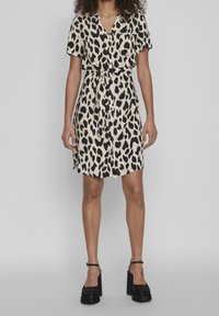 Robe à imprimé léopard beige et noir, manches courtes, encolure en V, taille ceinturée, longueur au genou, tissu texturé, associée à des escarpins noirs à talons carrés.