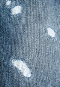 Denim azul desteñido con varios parches desgastados que exponen la tela blanca de abajo, con bordes deshilachados y una superficie texturizada.