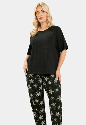 Donna bionda che indossa una maglietta nera a maniche corte larga con taschino sul petto e pantaloni neri decorati con motivi di stelle bianche.