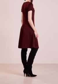 Vestido ajustado burdeos con mangas cortas, tejido texturizado, dobladillo acampanado, combinado con botas de tacón alto negras hasta el muslo, visto desde la parte posterior.