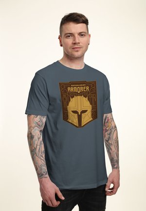 Homme tatoué portant un t-shirt bleu-gris avec un casque d'armure Mandalorien doré et le texte "Mandalorian Armorer" sur la poitrine.