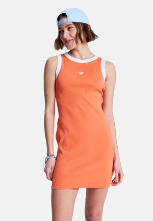 Junge Frau trägt ein oranges ärmelloses Kleid mit weißem Besatz und eine hellblaue Baseballkappe, die nach hinten getragen wird, lächelt und schaut leicht nach links.