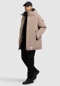 Chaqueta acolchada beige con capucha, cierre frontal, dos bolsillos laterales y parche con logo. Lleva encima un suéter negro y combinado con pantalones negros y botas.