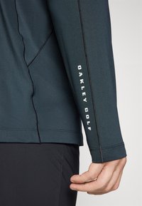 Mørk teal langermet golf skjorte med svart søm, med «OAKLEY GOLF» trykket i hvitt langs ermet. Strukturt stoff.
