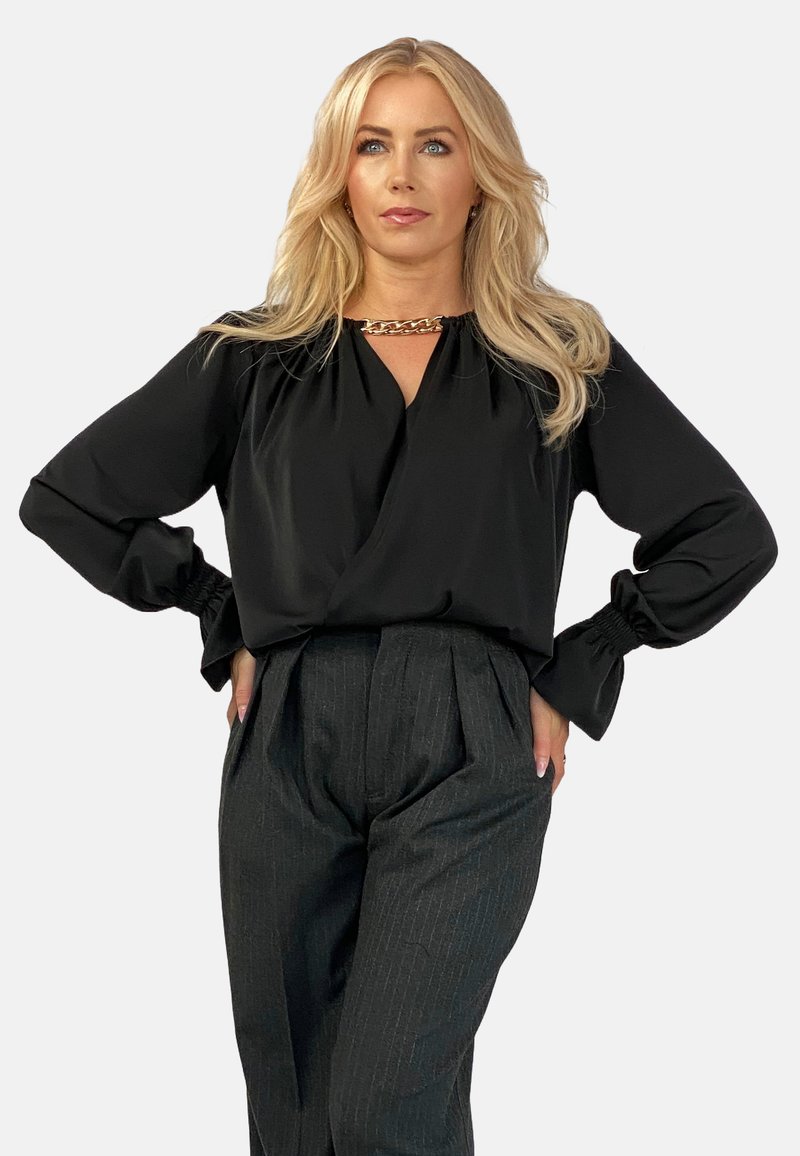 Czarna bluzka z długimi rękawami i marszczonymi mankietami, z głębokim dekoltem w kształcie litery V oraz złotym akcentem w postaci łańcuszka. W połączeniu z szarymi spodniami w prążki.