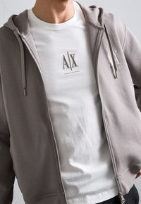 Felpa con cappuccio grigia con zip, orlo elastico e cappuccio con coulisse, sovrapposta a una t-shirt bianca con logo ricamato. Tessuto dalla texture liscia.