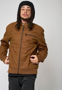Veste en cuir marron avec fermeture éclair sur le devant, poches latérales et poignets côtelés. Le design avec capuche présente des détails perforés sur les épaules.