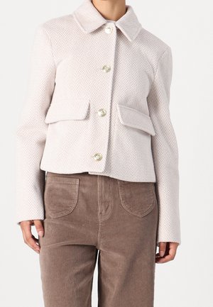 Vrouw die een lichtroze jasje met visgraatpatroon draagt, met knopen en klepzakken, gecombineerd met een hooggetailleerde bruine ribfluwelen broek.