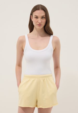 Jonge vrouw met lang bruin haar, een wit hemdje en gele shorts met een elastische taille dragend, staand met handen in de zakken.
