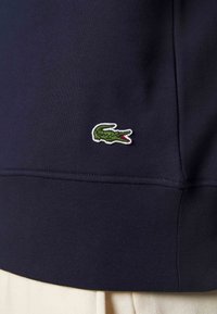 Section de tissu bleu marine foncé d'un vêtement avec un petit logo de crocodile vert brodé près de la couture au-dessus d'un pantalon de couleur claire.