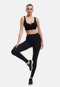 Czarny stanik sportowy z dekoltem w serek i szerokimi ramiączkami, w po�łączeniu z czarnymi legginsami o wysokim stanie. Model nosi czarne buty sportowe.