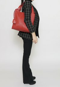 Rode leren tote bag met een gestructureerde afwerking, ontworpen met dubbele handvatten en een hexagonale label, weergegeven tegen een neutrale achtergrond.