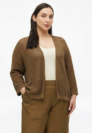 Frau mit dunklem Haar, die eine braune offen gestrickte Strickjacke, ein beiges Top und braune Hosen trägt, mit teilweise in den Taschen befindlichen Händen, vor weißem Hintergrund.