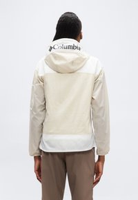 Άτομο που φοράει μπουφάν Columbia σε εκρού χρώμα με κουκούλα ανασηκωμένη, φαίνεται από πίσω, συνδυασμένο με καφέ παντελόνια πάνω σε απλό φόντο.