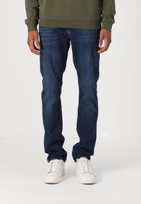 Mørkeblå denimjeans med klassisk passform, brukt med en grønn sweatshirt og hvite joggesko. Stoffet ser jevnt ut med lett tekstur.