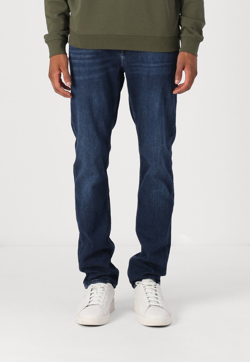 Mørkeblå denimjeans med klassisk passform, brukt med en grønn sweatshirt og hvite joggesko. Stoffet ser jevnt ut med lett tekstur.