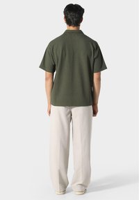 Lyhythihainen, tummanvihreä kauluspaita, jossa on teksturoitu kuosi, yhdistettynä vaaleanbeigeihin leveälahkeisiin housuihin ja rentoihin kenkiin.